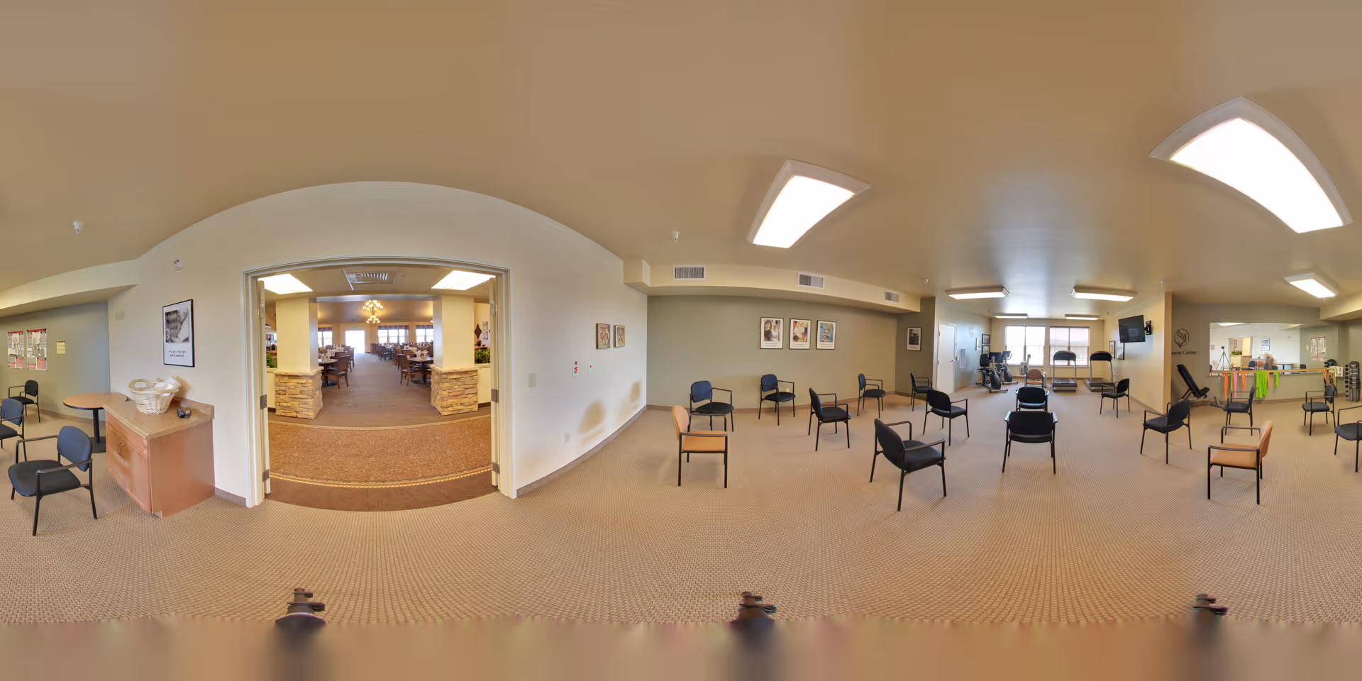 A spacious common area in a senior living facility with multiple chairs arranged in a circle, exercise equipment near the windows, and a view into a dining room through a wide doorway. The room has beige walls, carpeted floors, and ceiling lights.