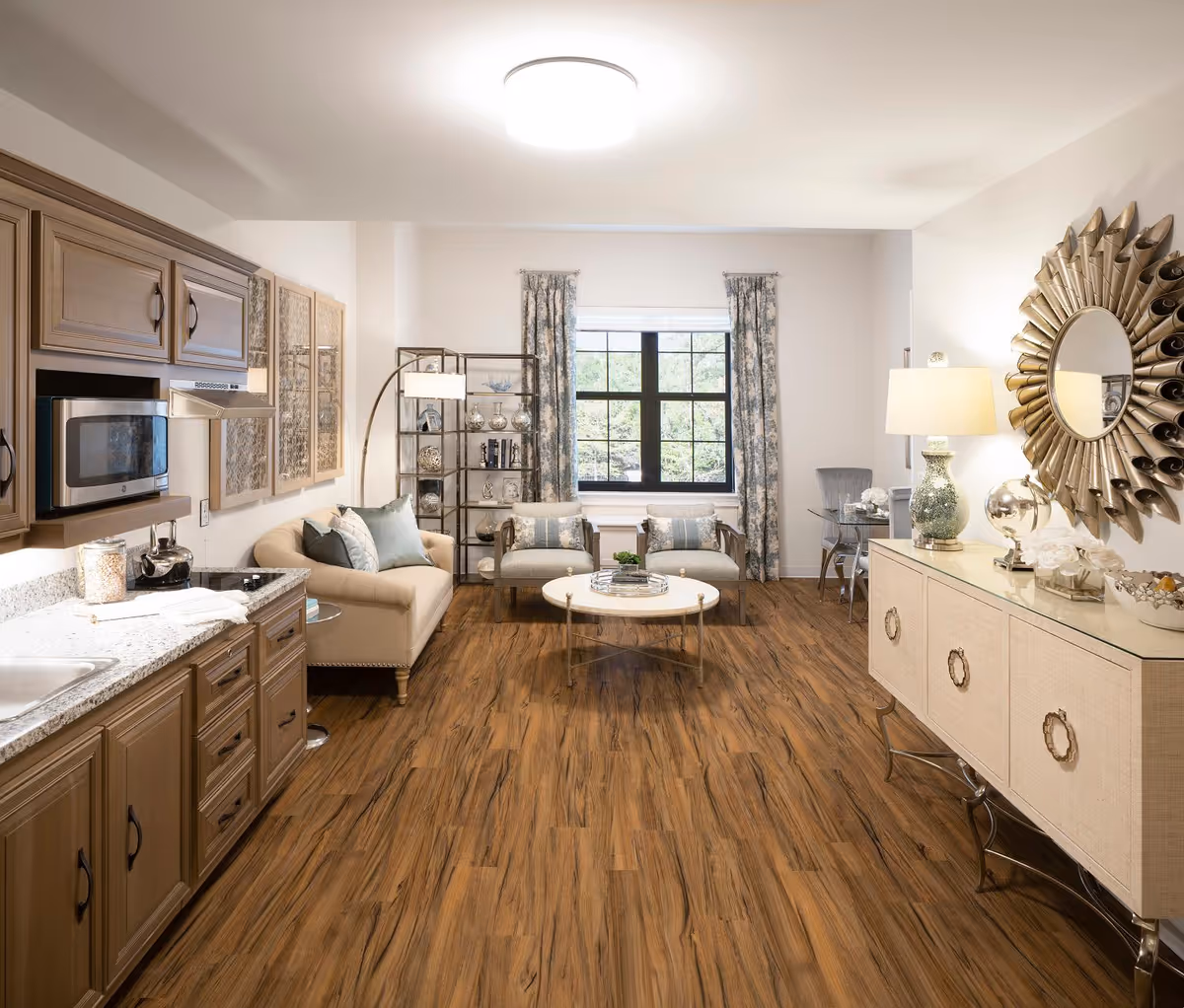 A bright and spacious senior living area featuring a small kitchen with wooden cabinets and granite countertops on the left, a beige sofa with blue and beige pillows, two armchairs, a round coffee table, and a large window with floral curtains. On the right side, there is a decorative sideboard with a lamp, a globe, and a large sunburst mirror above it. The room has wood flooring and a modern ceiling light.