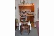 View of a small dining area with a wooden table covered by a white tablecloth and surrounded by wooden chairs. In the background, there is a kitchen with wooden cabinets, a stove, and various kitchen items on the counters and walls. A chandelier hangs above the dining table.