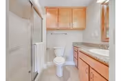 A clean bathroom featuring a glass-enclosed shower on the left, a white toilet in the center, and a wooden vanity with a granite countertop and sink on the right. There are wooden cabinets above the toilet and a mirror above the sink.