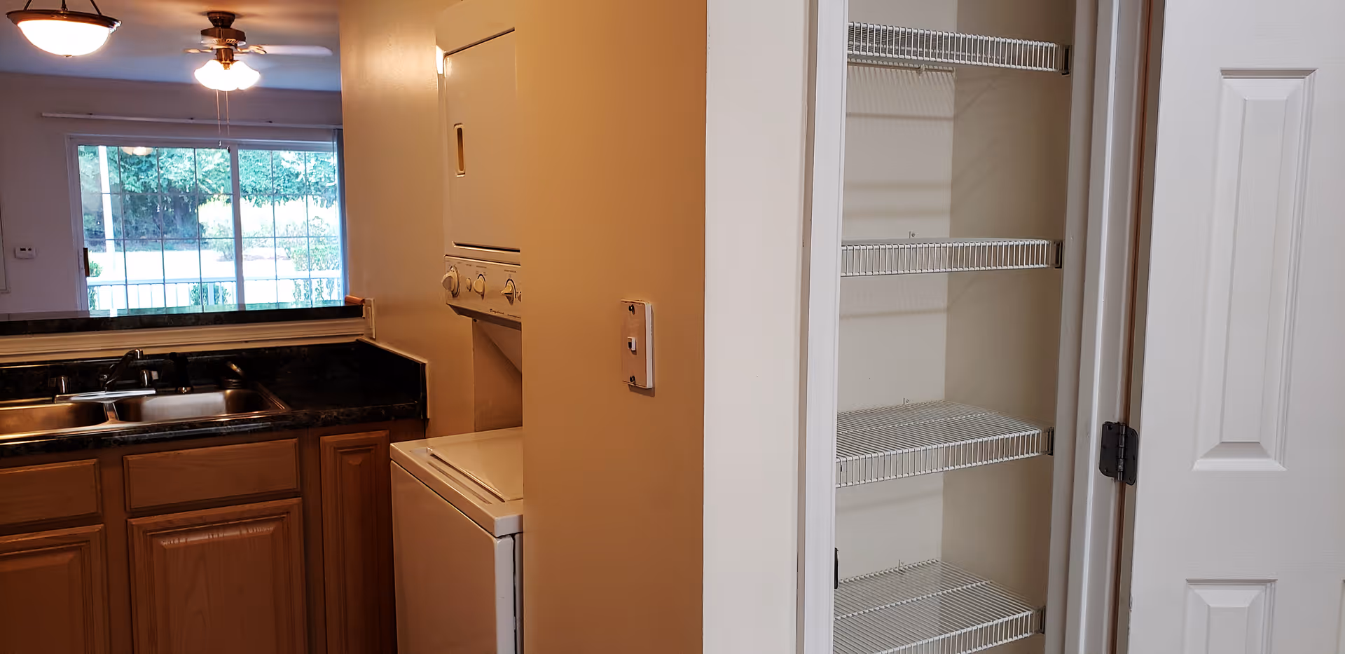Interior view of a kitchen area featuring a double sink with a dark countertop, wooden cabinets below, and a stacked washer and dryer unit next to a pantry with wire shelving. A large window in the background lets in natural light, and ceiling lights are visible.