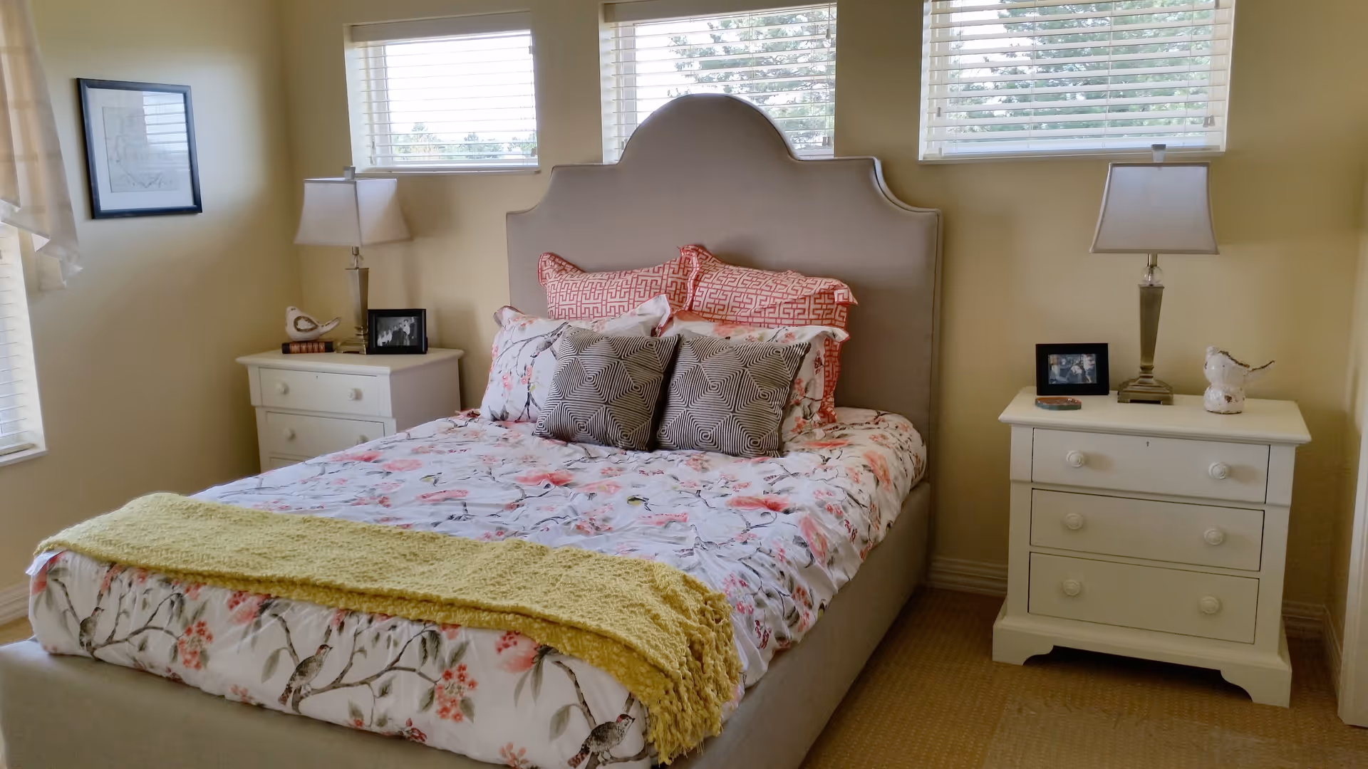 A cozy bedroom with a beige upholstered bed featuring floral and patterned pillows and a floral bedspread. There is a yellow knitted throw blanket at the foot of the bed. On each side of the bed are white nightstands with table lamps, framed photos, and decorative bird figurines. Three windows with white blinds are above the bed, letting in natural light.