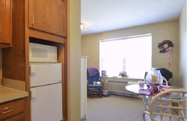 A small kitchen area with a white microwave and refrigerator built into wooden cabinetry on the left, leading into a bright living room with a large window. The living room has a blue cushioned rocking chair with a purple blanket, a round glass-top table with decorative items, and a wall-mounted air conditioning unit below the window. A floral wreath with a pink ribbon hangs on the right wall.
