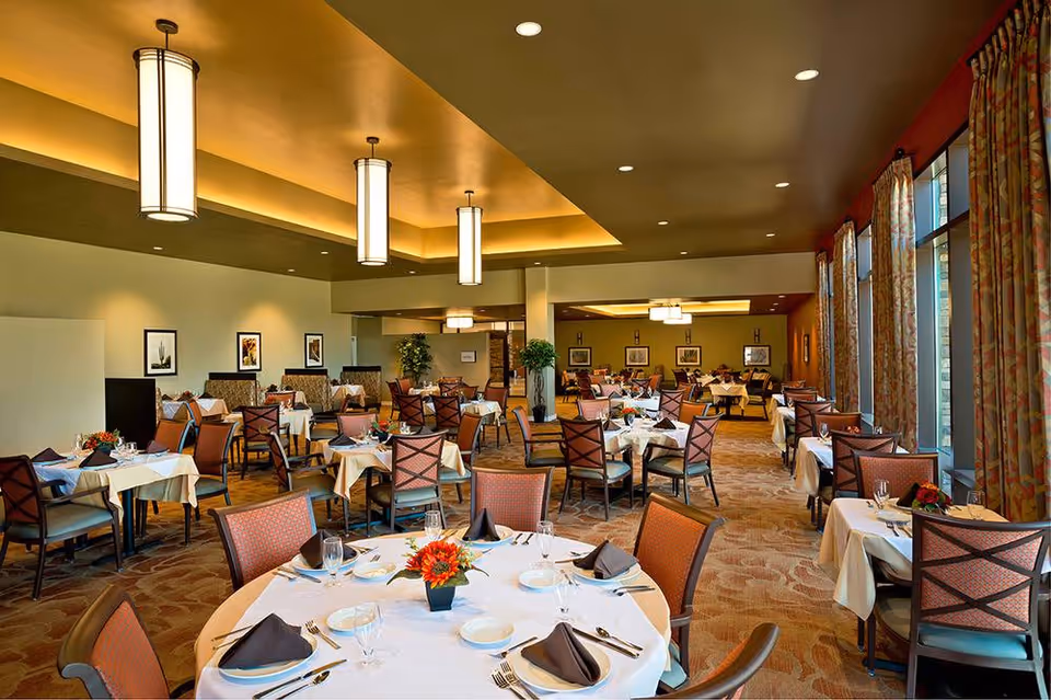 A spacious dining room with multiple round and square tables covered with white tablecloths, each set with plates, glasses, silverware, and napkins. The room has warm lighting from ceiling fixtures and large windows with patterned curtains on the right side. There are framed pictures on the walls and some potted plants adding greenery to the space.