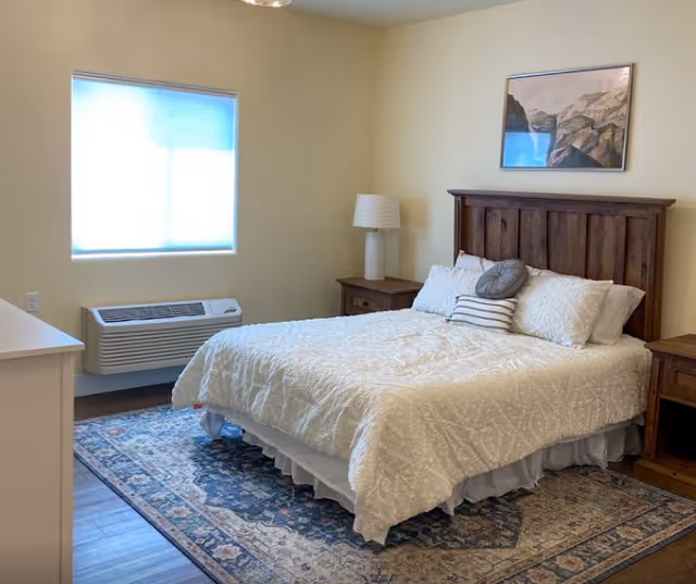 Sunlit furnished bedroom with a bed featuring a wooden headboard, two nightstands, a lamp, area rug, and a window unit.