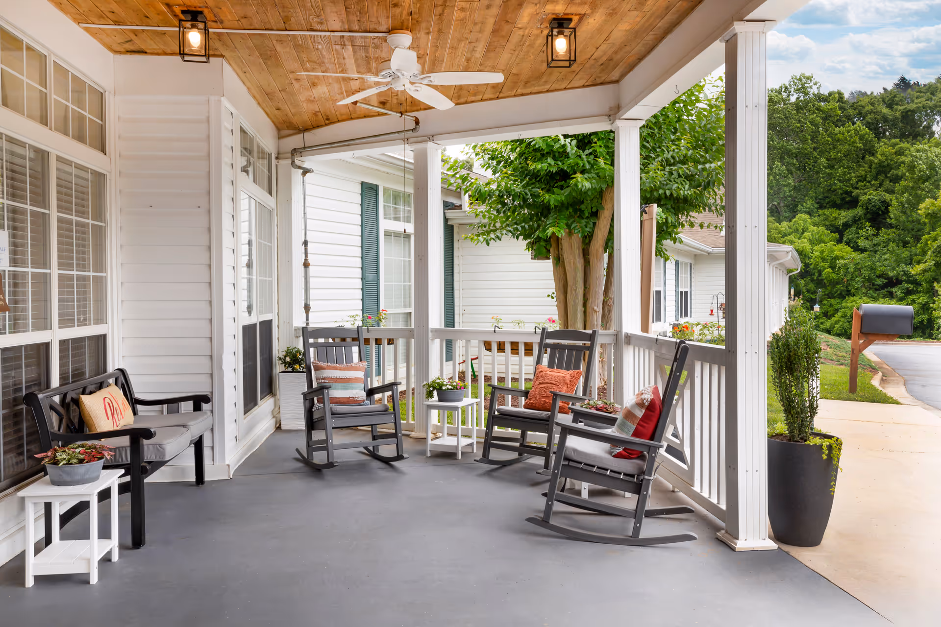Covered porch area with a wooden ceiling and white ceiling fan, featuring three gray rocking chairs with colorful cushions, a black bench with a cushion and a pillow, small white side tables with potted plants, white railing, and a view of greenery and a mailbox near a paved driveway.