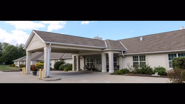 Exterior view of a single-story senior living facility building with a covered entrance supported by columns, surrounded by shrubs and greenery under a blue sky with some clouds.