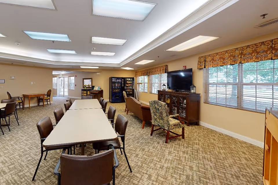 A spacious senior living common area with a long table surrounded by chairs in the center, additional seating including armchairs and a floral-patterned chair near a large flat-screen TV on a wooden cabinet. The room has large windows with patterned valances, beige walls, and carpeted flooring. In the background, there is a vending machine and a small table with chairs near the entrance.