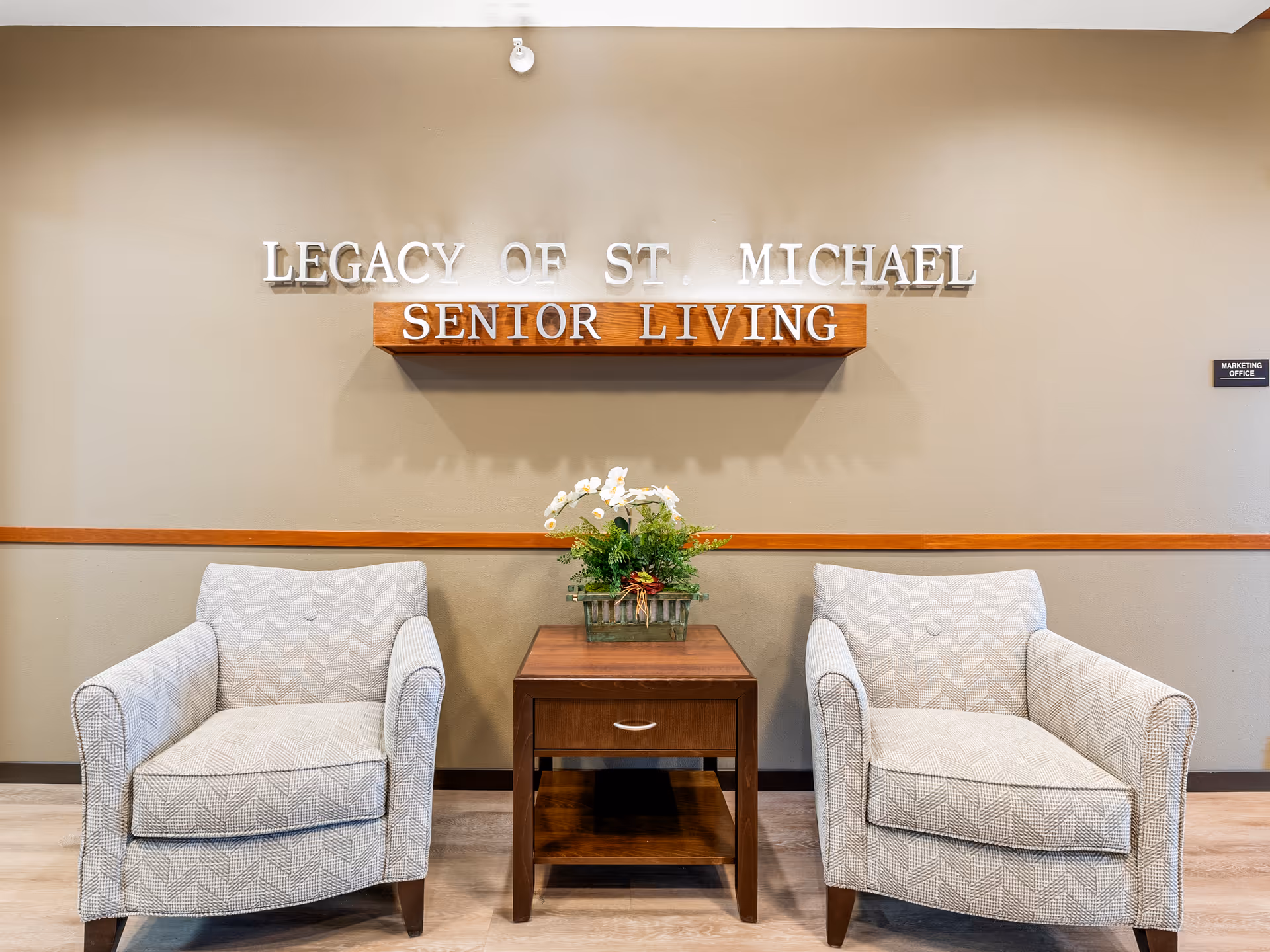 Two patterned armchairs with a wooden side table between them holding a flower arrangement, set against a beige wall with the sign 'Legacy of St. Michael Senior Living' mounted above.