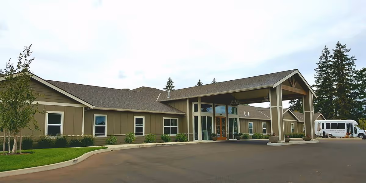 Exterior view of Cherrywood Memory Care facility showing a single-story building with a covered entrance, several windows, landscaped greenery, and a white shuttle bus parked near the entrance.