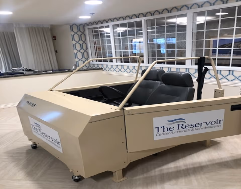 A beige driving simulator setup with two black seats and a steering wheel, placed indoors in a room with patterned wallpaper and large windows. The simulator has signs on the side and front that read 'The Reservoir Center for Health & Rehabilitation.'