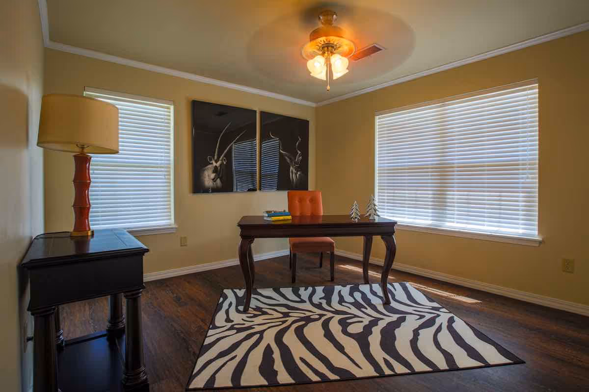 A small office or study room with a wooden desk and an orange chair placed on a zebra-striped rug. There is a side table with a lamp on the left, two windows with blinds, and two black and white animal-themed artworks on the wall. A ceiling fan with lights is mounted on the ceiling.