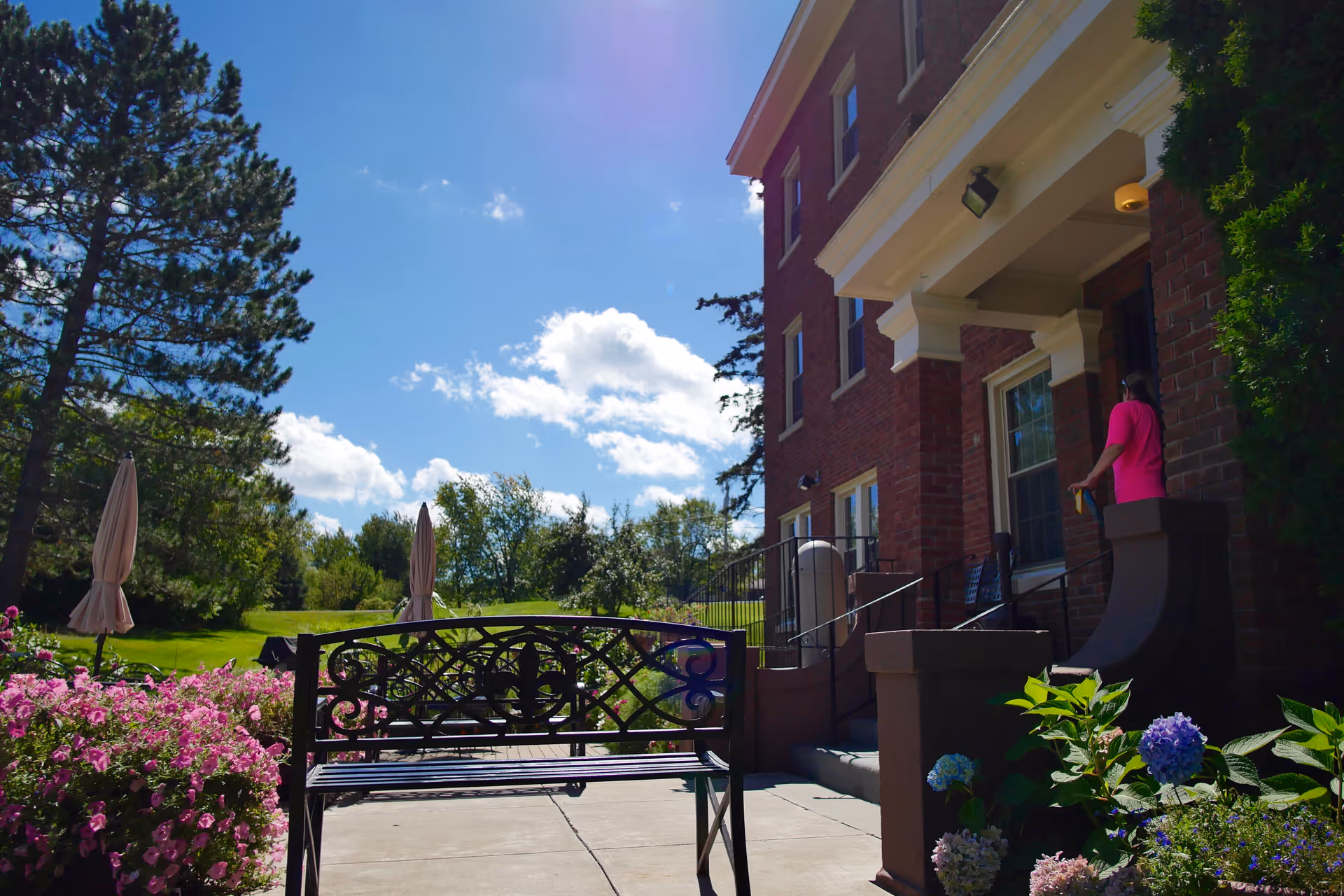 Outdoor patio area at McCarthy Manor with a decorative metal bench in the foreground, colorful flowers and shrubs on either side, and a brick building entrance with a person standing near the door. The sky is blue with some clouds and there are trees and green grass in the background.