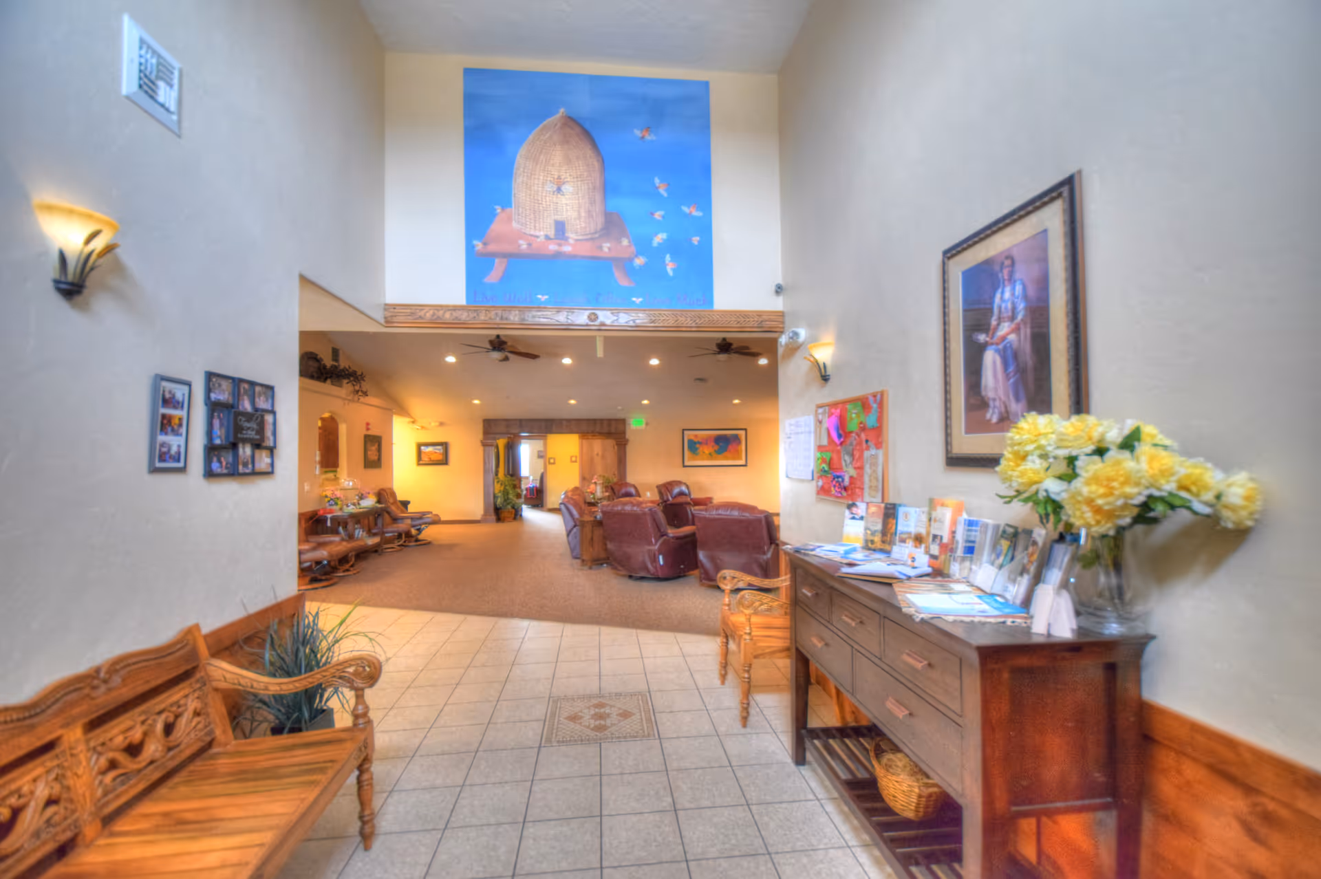 Interior view of a senior living facility hallway leading to a common area with leather chairs. The hallway has tiled flooring and wooden benches along the walls. On the right side, there is a wooden console table with brochures and a vase of yellow flowers. The walls are decorated with framed pictures and a large painting of a beehive with bees on the far wall above the entrance to the common area.