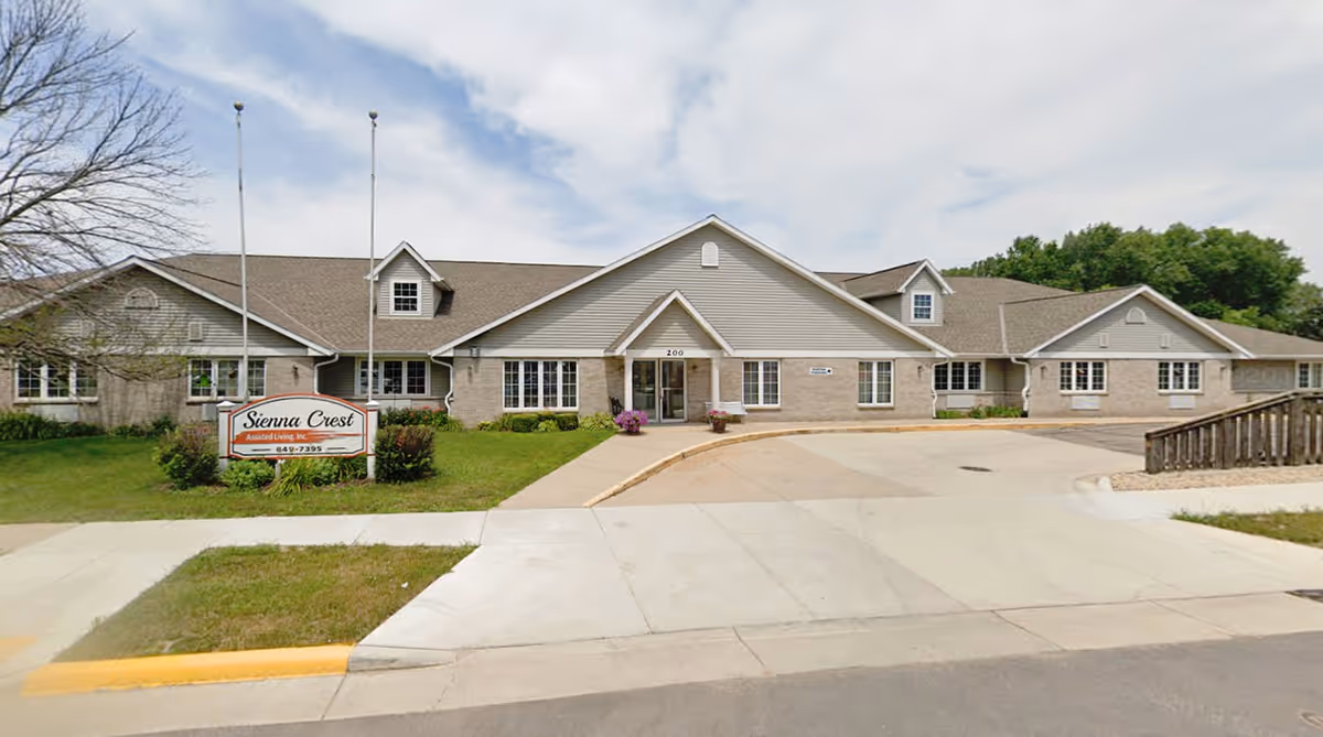 Front exterior view of Sienna Crest Assisted Living, Inc., a single-story building with beige siding and brick accents, a peaked roof, and multiple windows. There is a sign on the lawn with the facility's name and phone number, two flagpoles without flags, a curved driveway, and some landscaping with grass and bushes.