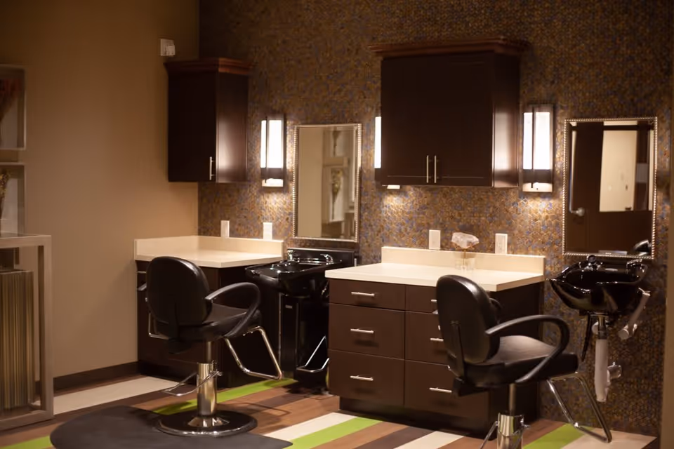 Interior view of a salon area with two black salon chairs in front of two black wash basins and mirrors mounted on a wall with patterned wallpaper. There are dark wood cabinets above and below the countertops, with wall-mounted lights beside the mirrors. The floor has a striped pattern with green, brown, and beige colors.