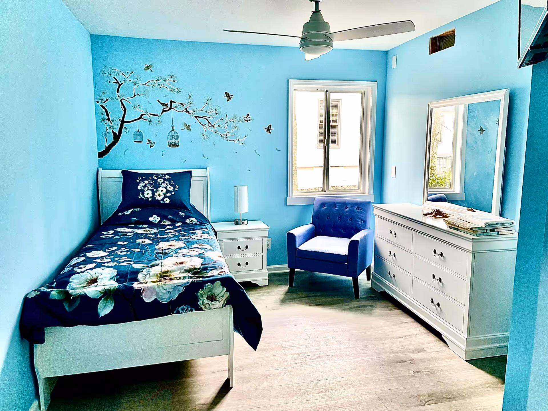A bright bedroom with light blue walls featuring a floral and bird mural above a white single bed with a dark blue floral bedspread. Next to the bed is a white nightstand with a lamp. Across from the bed is a white dresser with a large mirror and some books on top. A blue upholstered armchair is positioned near a window letting in natural light. The room has light wood flooring and a ceiling fan.