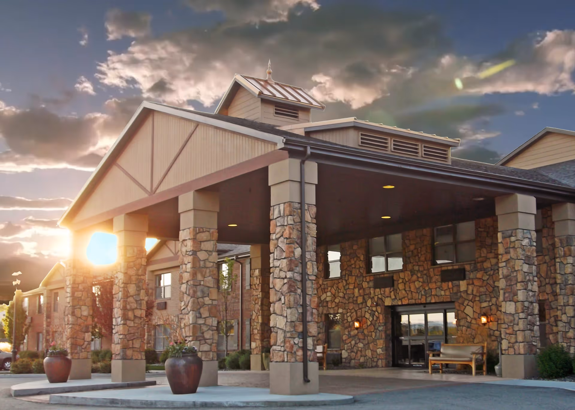 Exterior view of a senior living facility entrance with stone pillars and a covered driveway at sunset, featuring a bench and potted plants near the entrance.
