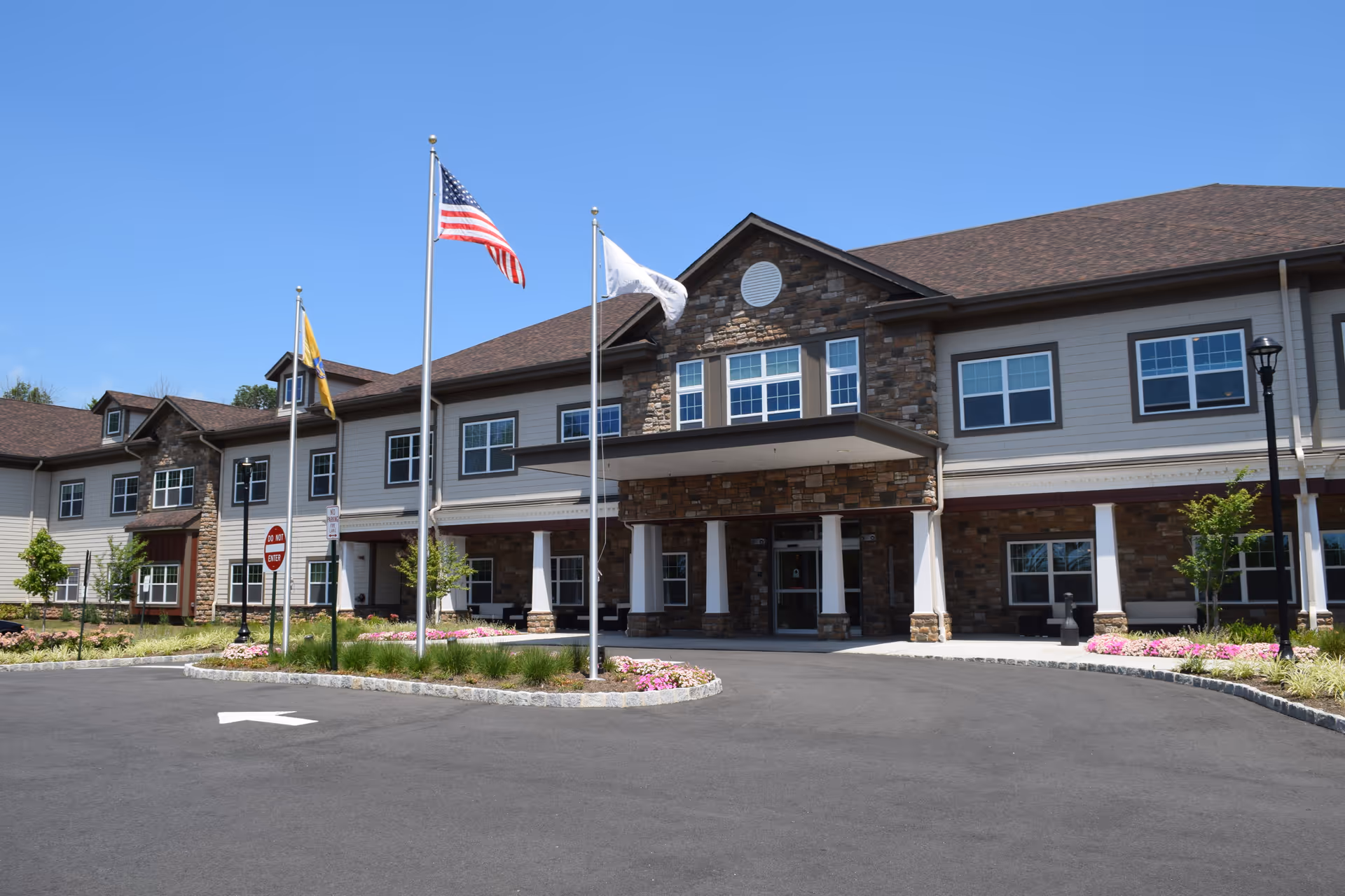 Front entrance of an assisted living building with flagpoles, a circular driveway, and landscaped flower beds.