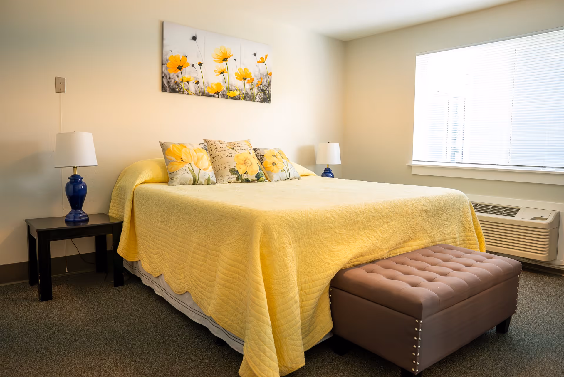 A bedroom with a large bed covered in a yellow quilt and three decorative pillows with floral designs. There are two blue lamps on black bedside tables on either side of the bed. A brown tufted bench sits at the foot of the bed. A window with blinds and an air conditioning unit is on the right wall. A wall art piece featuring yellow flowers hangs above the bed.
