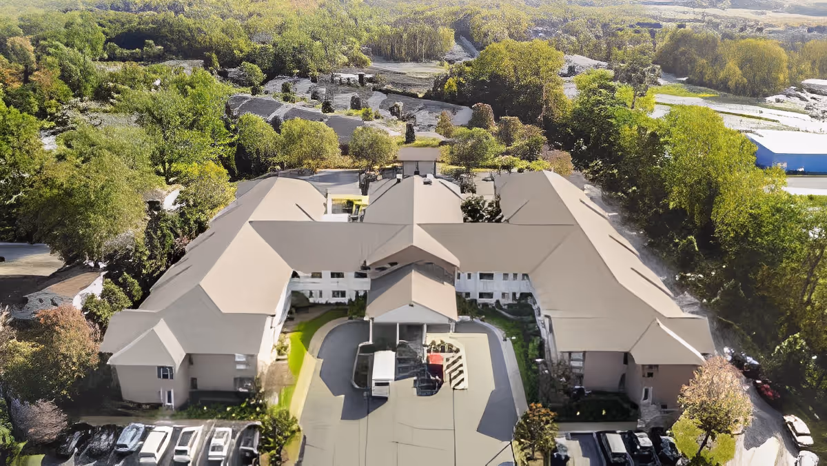 Aerial view of a large senior living facility building surrounded by trees and greenery, with a parking area in front and a driveway leading to the main entrance.