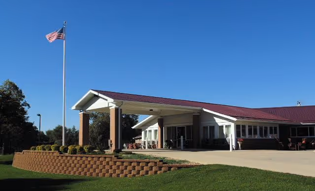 Front entrance of a single-story convalescent home with a covered drive, flagpole, and landscaped lawn under a clear blue sky.