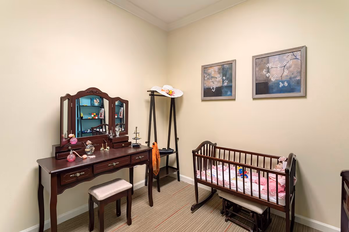 A small room with beige walls and carpeted floor featuring a dark wooden vanity table with a tri-fold mirror and a cushioned stool. On the right side, there is a wooden baby crib with pink bedding and toys inside. A black coat rack stands between the vanity and the crib, holding a white hat and an orange scarf. Two framed floral paintings hang on the wall above the crib.
