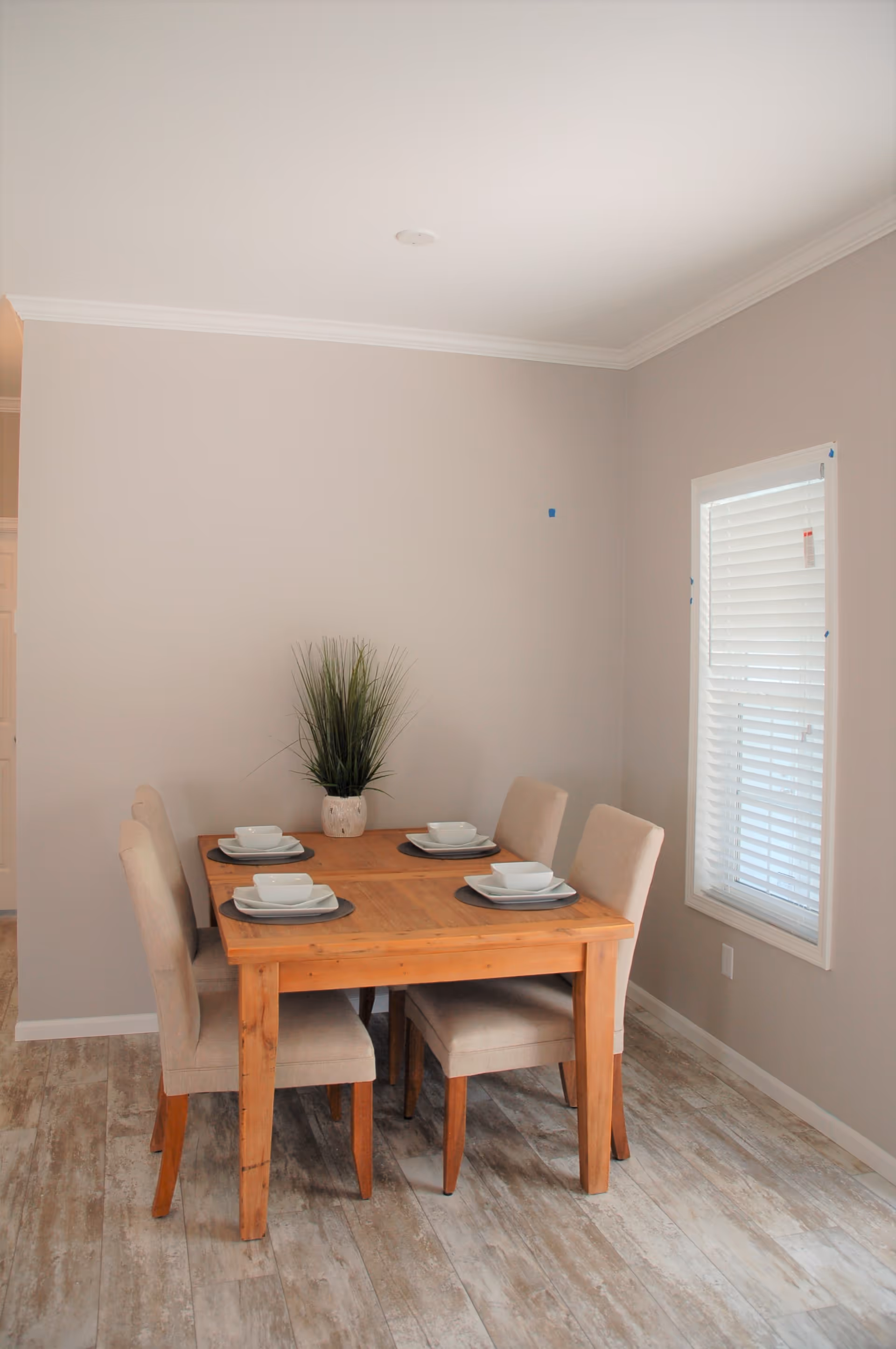 A small dining area with a wooden table set for four people. The table has four beige upholstered chairs and is set with white square plates, bowls, and cups on dark placemats. A potted green plant is placed in the center of the table. The room has light-colored walls, a window with white blinds, and wood-look flooring.