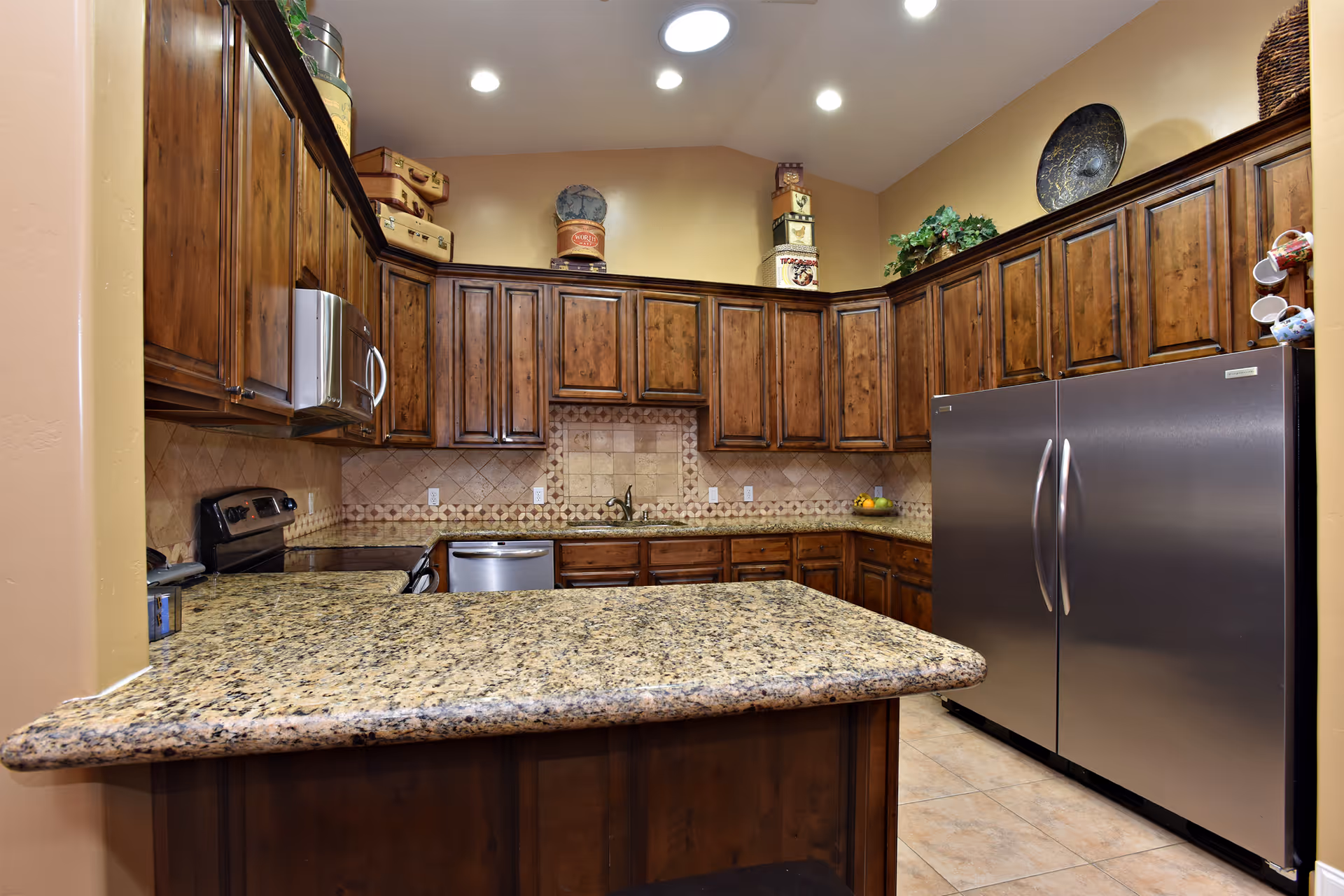 Spacious kitchen with a granite island, dark wooden cabinets, and stainless steel appliances including a double-door refrigerator.