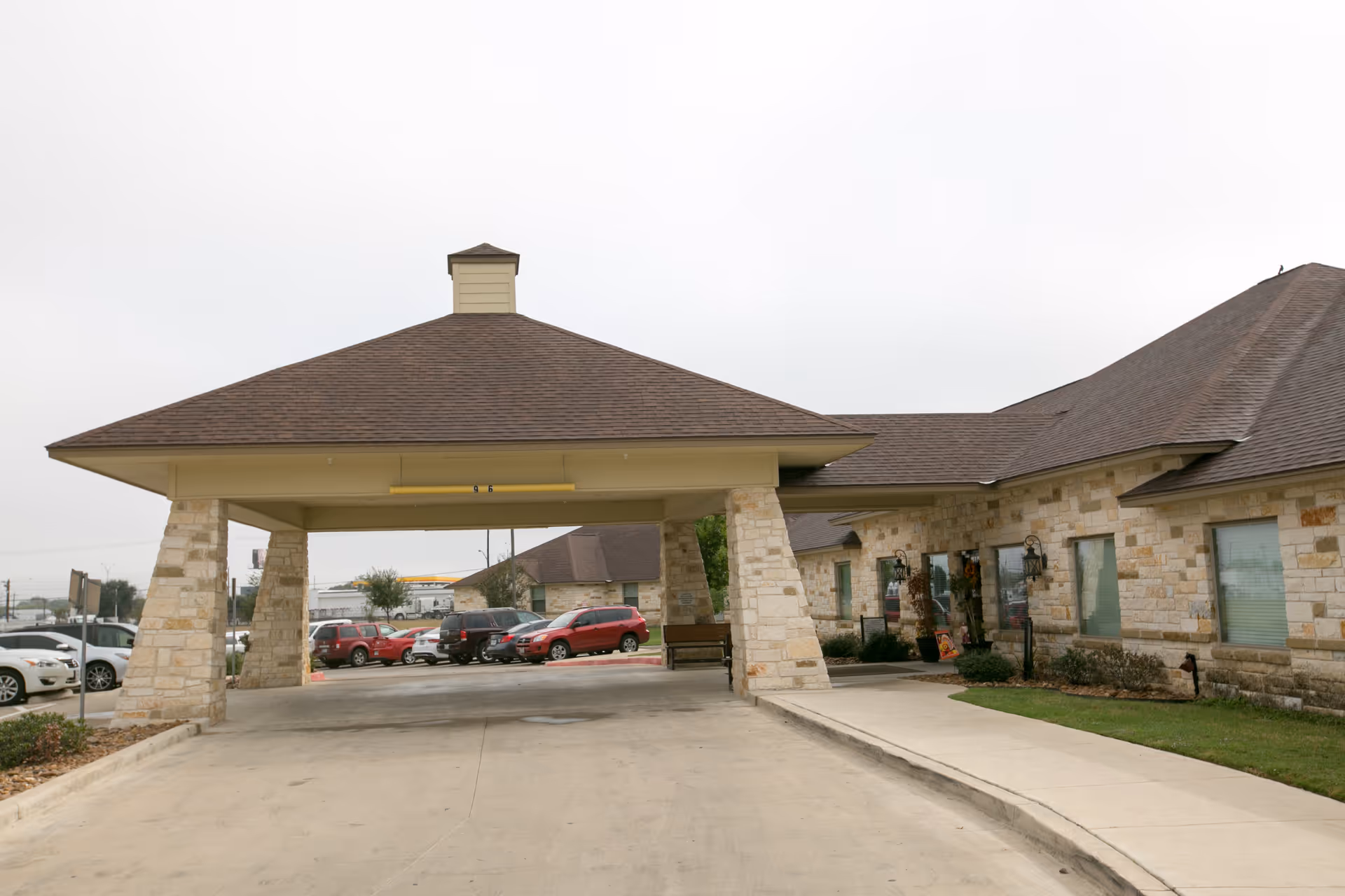 Entrance area of Legend Oaks Healthcare and Rehabilitation - West San Antonio showing a covered driveway with stone pillars and a building with stone exterior walls and multiple windows. Several parked cars are visible in the background under an overcast sky.