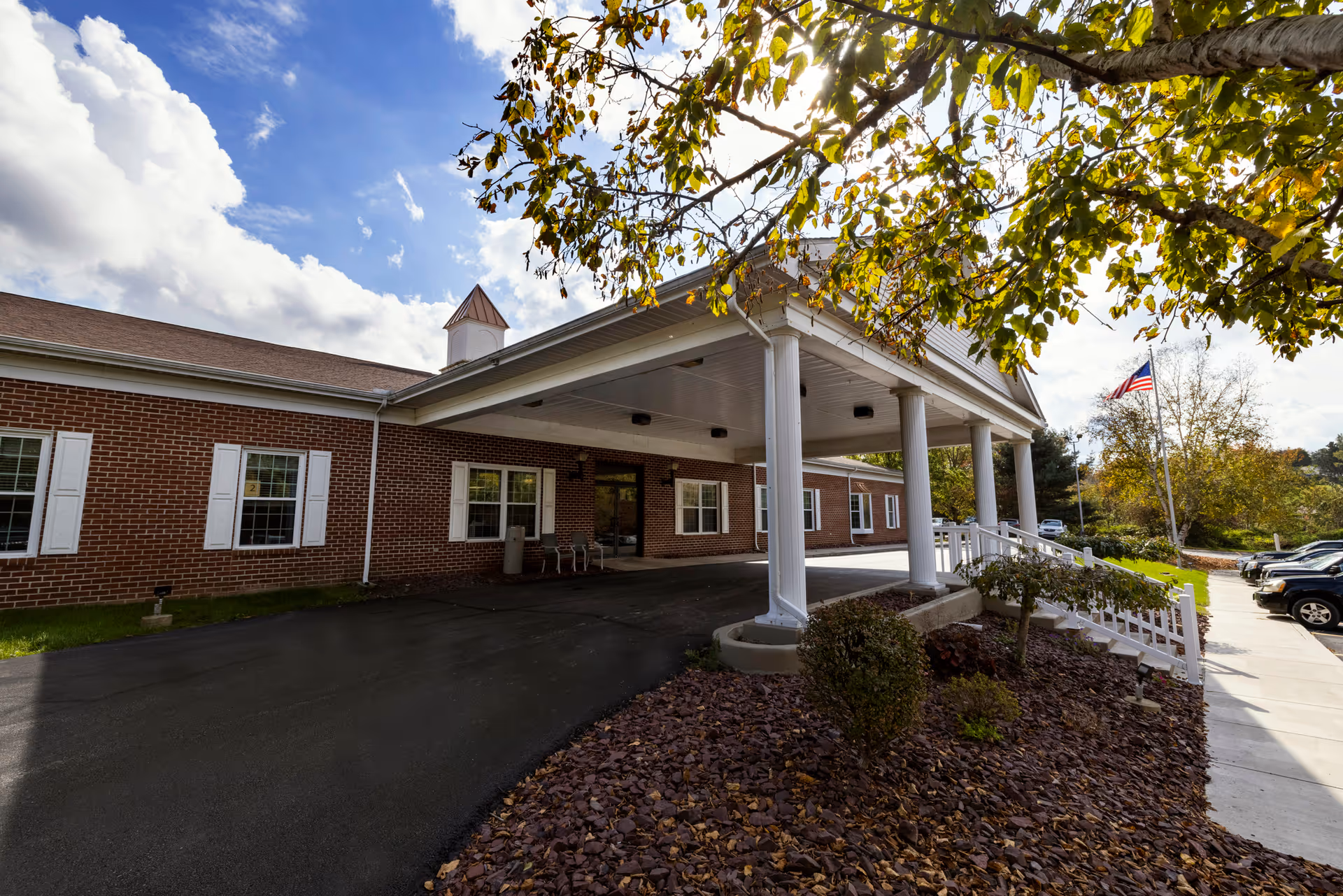 Front entrance of a brick senior living facility with a covered portico supported by white columns, landscaping, and an American flag.