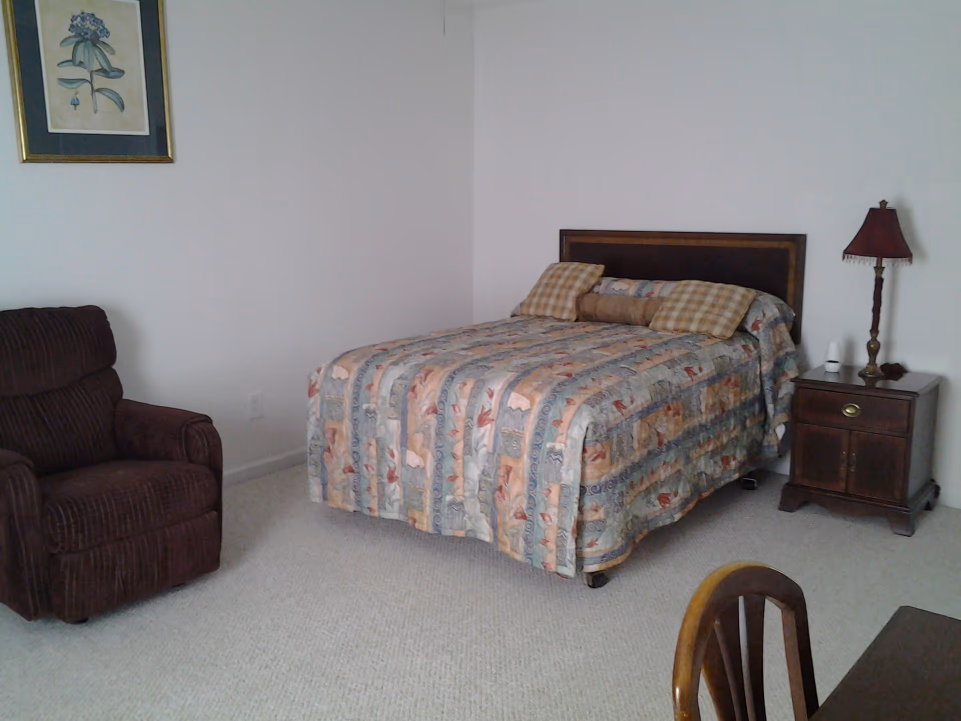 A bedroom with a bed covered in a patterned quilt, two plaid pillows, a brown armchair, a wooden nightstand with a red lamp and a small white device, a framed botanical print on the wall, and a wooden chair partially visible in the foreground.