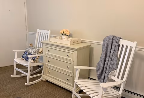 Two white wooden rocking chairs flanking a small white three-drawer dresser topped with a tray and flowers in a simple interior seating area.