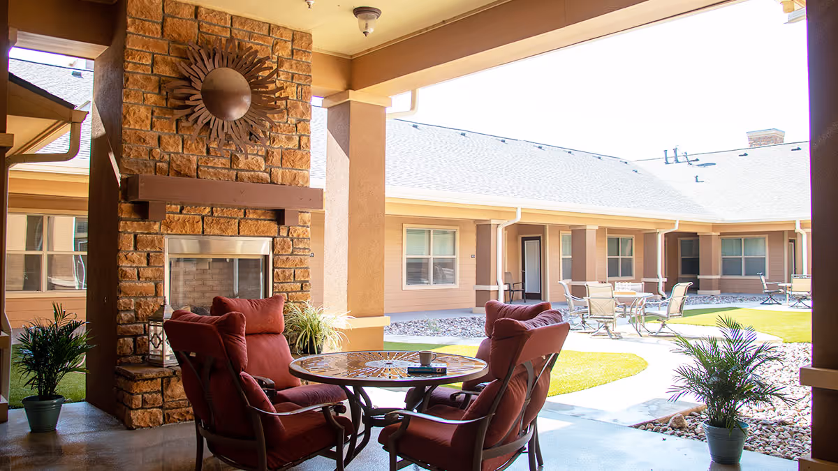 Covered courtyard seating area with red cushioned chairs around a round table by a stone fireplace, overlooking a sunny central courtyard with patio tables.