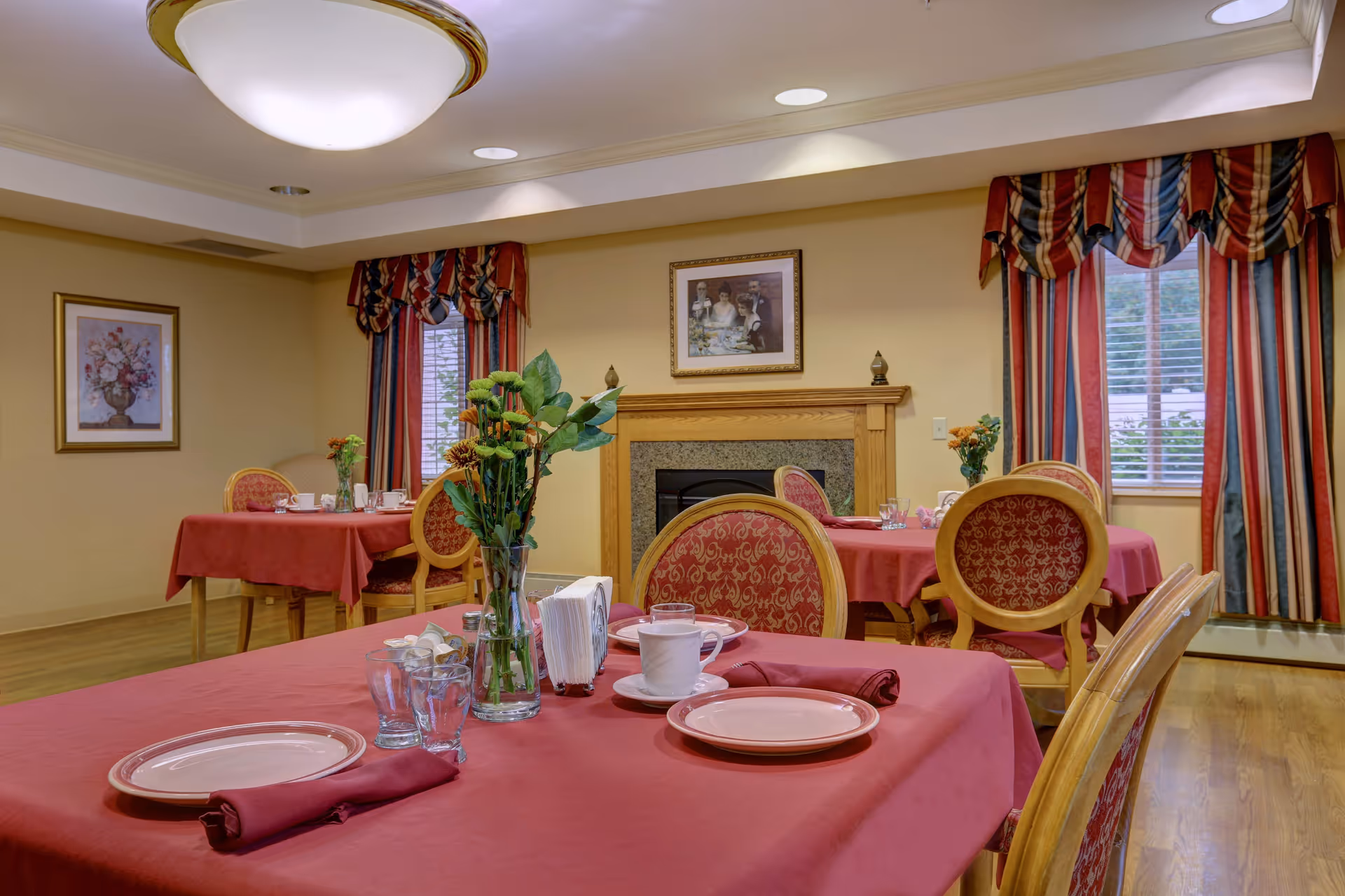Dining room with red-tableclothed tables, floral centerpieces and upholstered chairs arranged near a fireplace and windows.