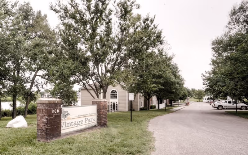Exterior view of Vintage Park At Waterfront senior living community showing a driveway lined with trees and parked cars, a brick sign with the community name, and beige buildings in the background.