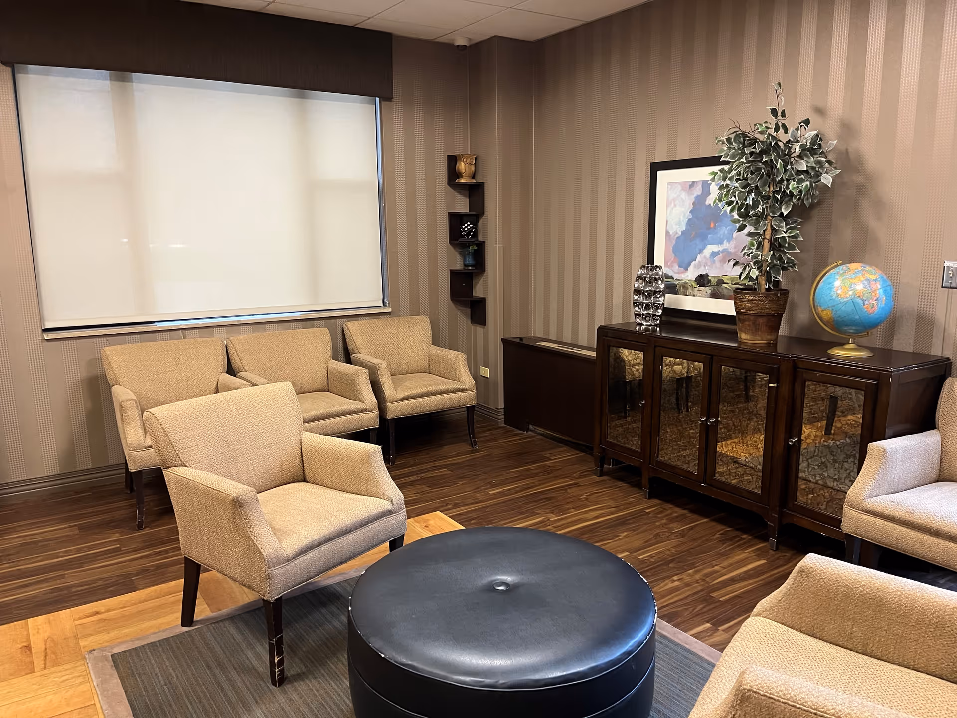 A cozy lounge with beige armchairs arranged around a round black ottoman, a wooden cabinet with decor and a globe, and a large window with a roller shade.