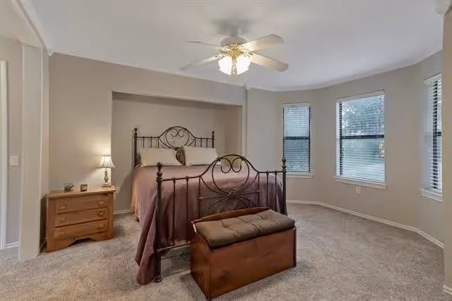 Light, carpeted bedroom with an ornamental iron bed dressed in purple bedding, a nightstand with lamp, ceiling fan, bay windows, and a storage bench at the foot of the bed.