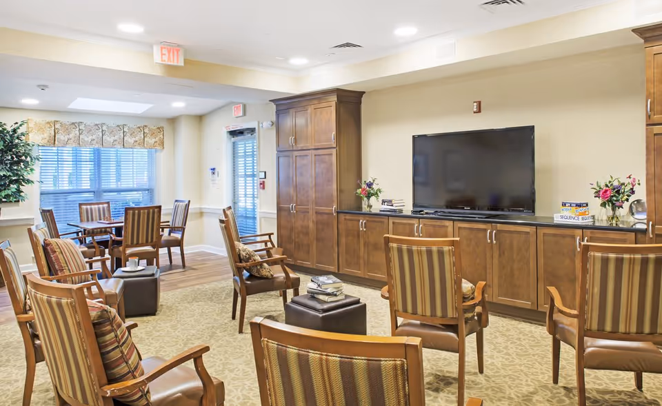 Community lounge with multiple striped armchairs arranged around a central ottoman facing a large flat-screen TV and wooden cabinetry.