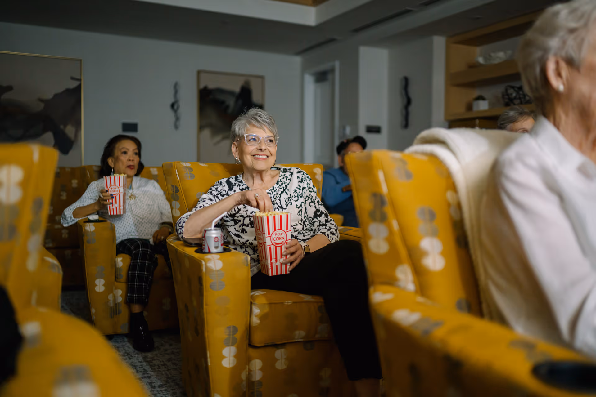 Several elderly people sitting in yellow patterned armchairs in a cozy room, enjoying popcorn and a canned drink, appearing to watch something with interest and smiles.
