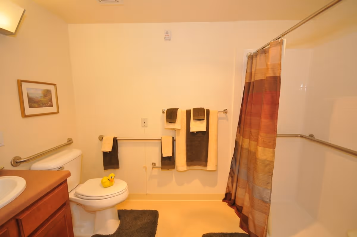 A bathroom with a toilet, sink, and shower area. The toilet has a yellow rubber duck on the seat. There are multiple towels hanging on metal bars on the wall, and a multicolored shower curtain is partially drawn. A framed picture hangs on the wall above the toilet, and there are dark-colored bath mats on the floor.