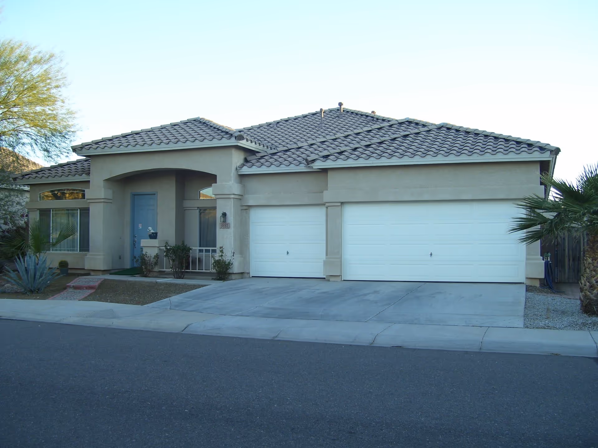 Front exterior view of a single-story house with a tiled roof, beige stucco walls, a blue front door, two large white garage doors, and some desert landscaping including palm trees and shrubs.