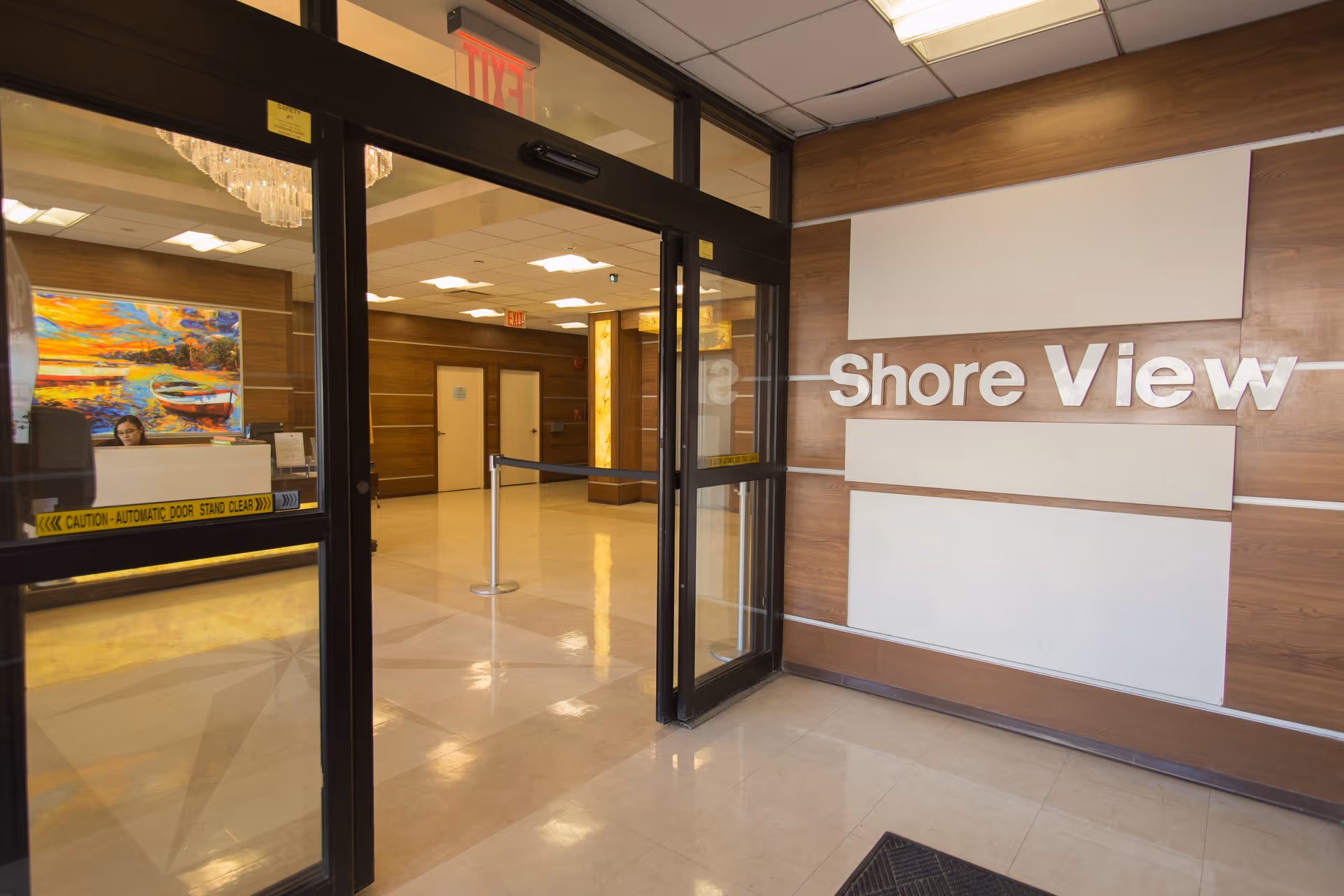 Entrance area of Shore View Nursing and Rehabilitation facility showing automatic glass doors, a reception desk with a person behind it, a colorful painting on the wall, and a sign on the wall that reads 'Shore View'.