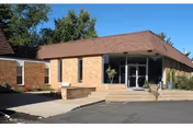 Exterior view of a single-story brick building with large windows and a sloped roof under a clear blue sky. There is a paved driveway and a small landscaped area with a tree and shrubs near the entrance.