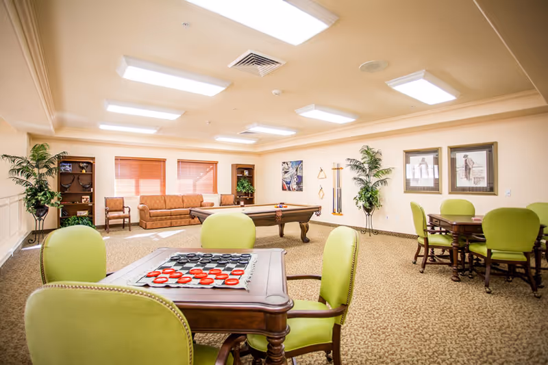 A bright and spacious senior living common area with beige walls and carpet. The room features a pool table in the center, a checkers game set up on a wooden table with green upholstered chairs in the foreground, and additional tables and chairs in the background. There are two large plants, framed artwork on the walls, and a sofa with chairs near windows with wooden blinds.