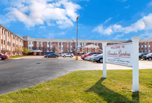 Exterior view of Brenden Gardens senior living facility showing a large brick building with multiple windows, a parking lot with several cars, a grassy area, and a white sign welcoming visitors to Brenden Gardens under a partly cloudy blue sky.