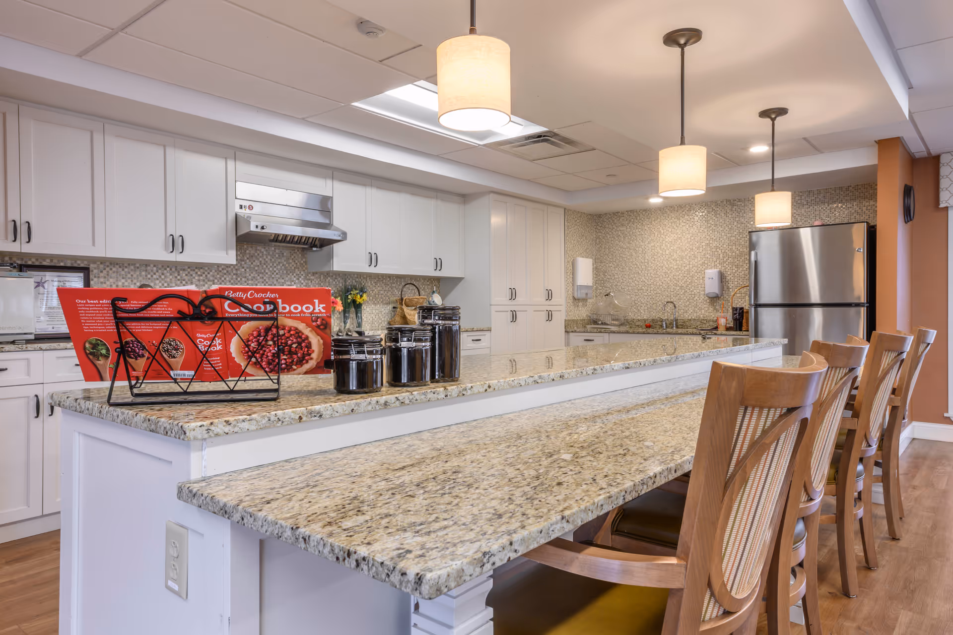 A modern kitchen with white cabinets, granite countertops, and a tiled backsplash. There is a long kitchen island with a raised bar area and wooden chairs lined up along it. On the countertop, there is a Betty Crocker cookbook in a stand and several black canisters. Stainless steel appliances, including a refrigerator and a range hood, are visible. The room is well-lit with ceiling lights and pendant lamps.