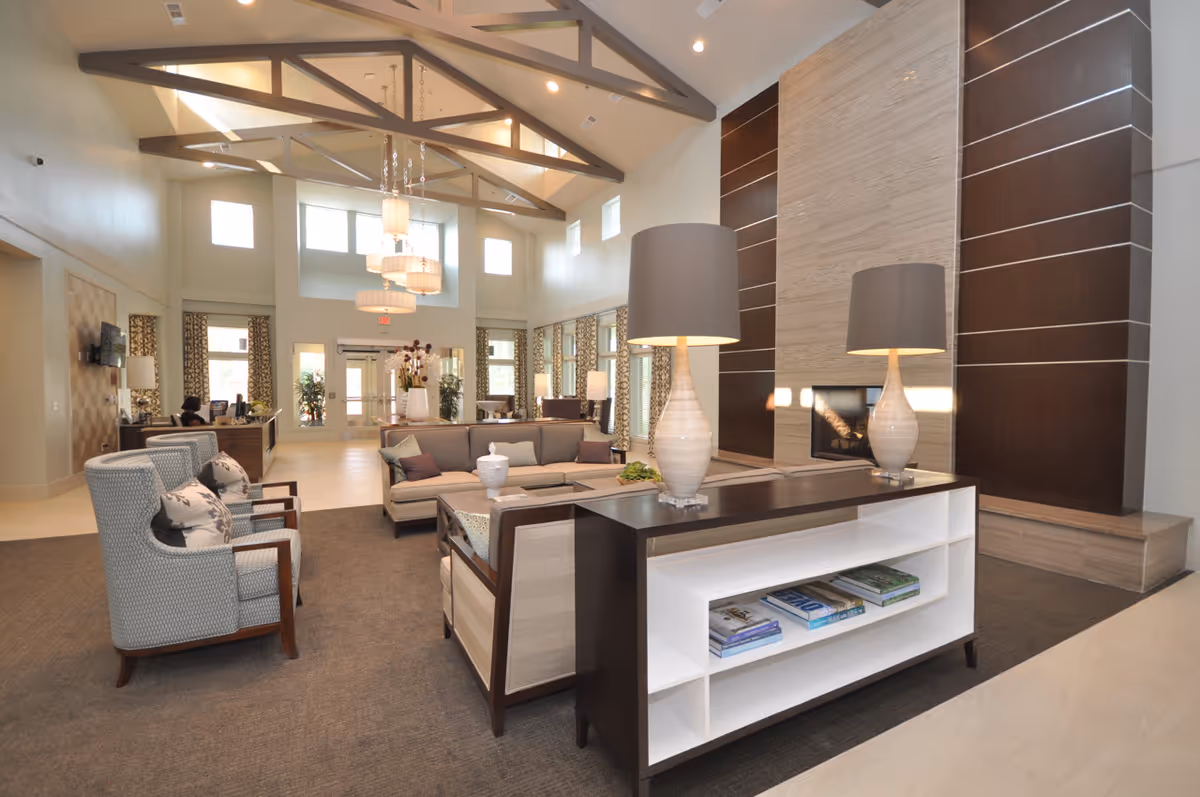 A spacious and well-lit living room area in The Crossings Retirement Community featuring modern furniture including armchairs and sofas arranged around a central table with decorative lamps. The room has high ceilings with exposed beams, large windows with patterned curtains, a tall fireplace with a stone surround, and a reception desk visible in the background.