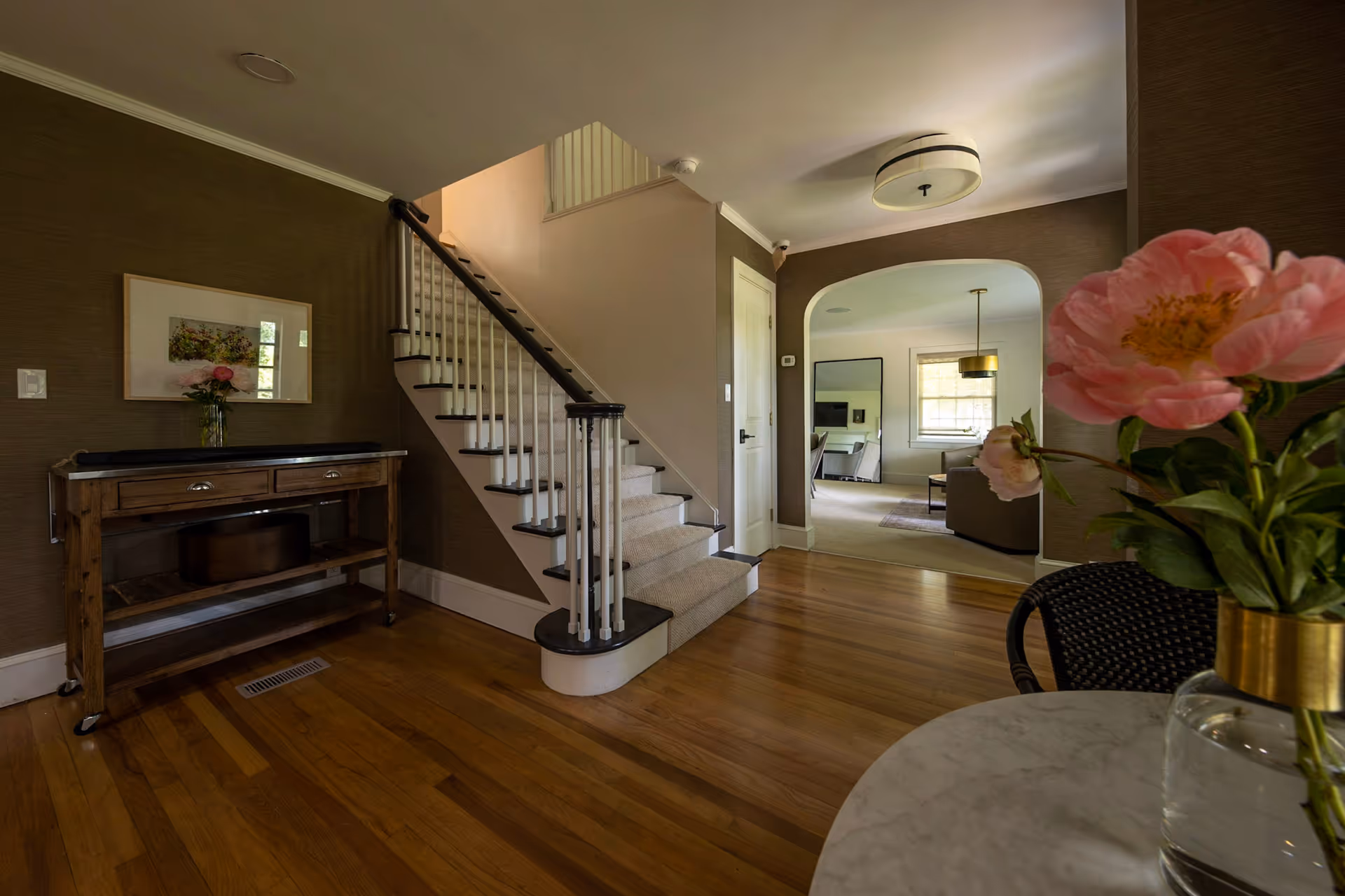 Interior view of a residential living space showing a wooden floor, a staircase with white railings and dark handrails, a wooden console table with a framed picture and a vase with flowers on it, and an archway leading to a living room area with a sofa, a large mirror, and a window with blinds. In the foreground, there is a round table with a glass vase holding pink flowers.