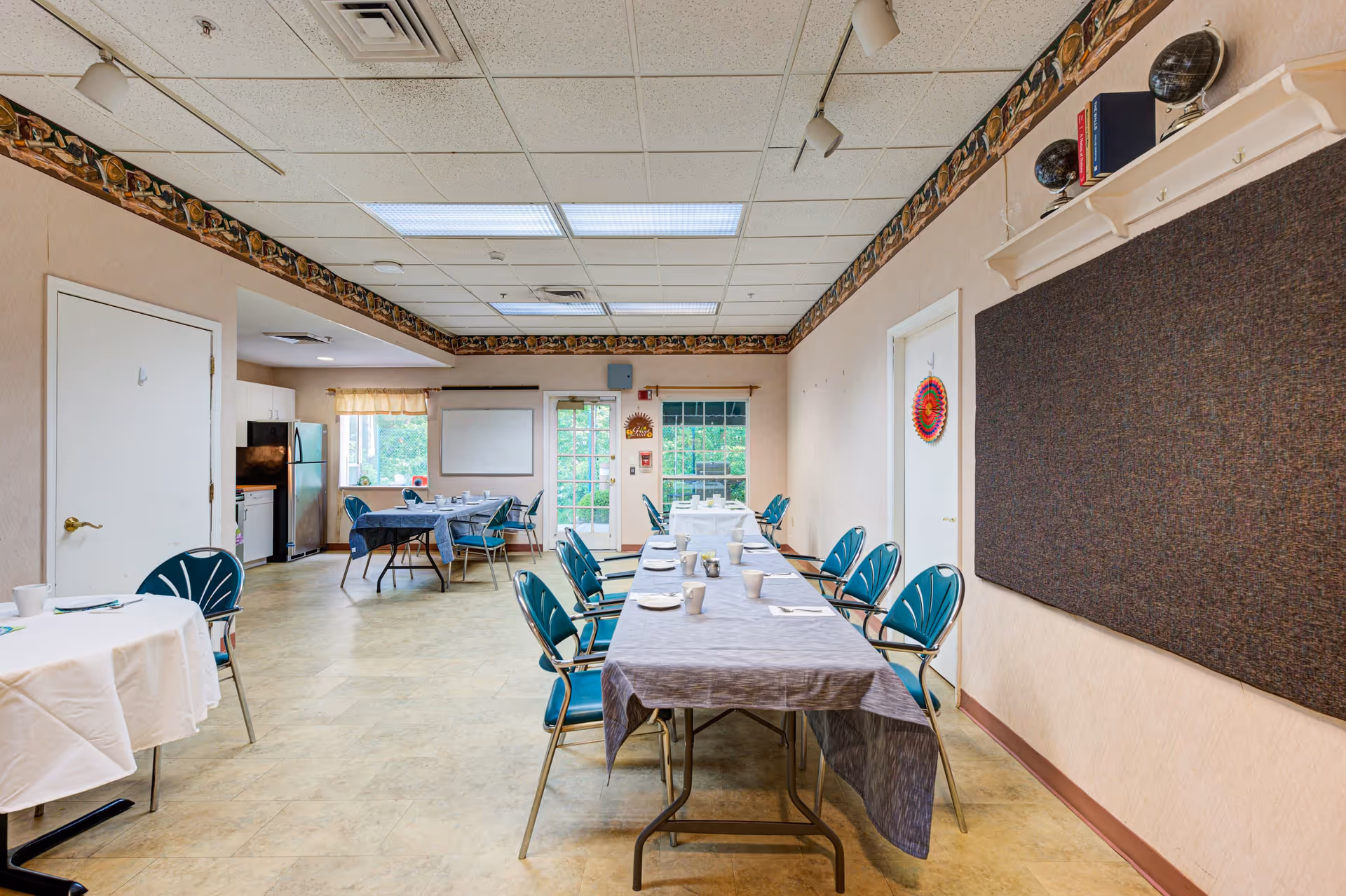 A dining room in a senior living facility with tables covered in tablecloths and set with cups, plates, and utensils. Blue chairs are arranged around the tables. The room has a beige floor and walls with a decorative border near the ceiling. There is a kitchen area with a refrigerator and cabinets visible in the background, and a door and windows letting in natural light.
